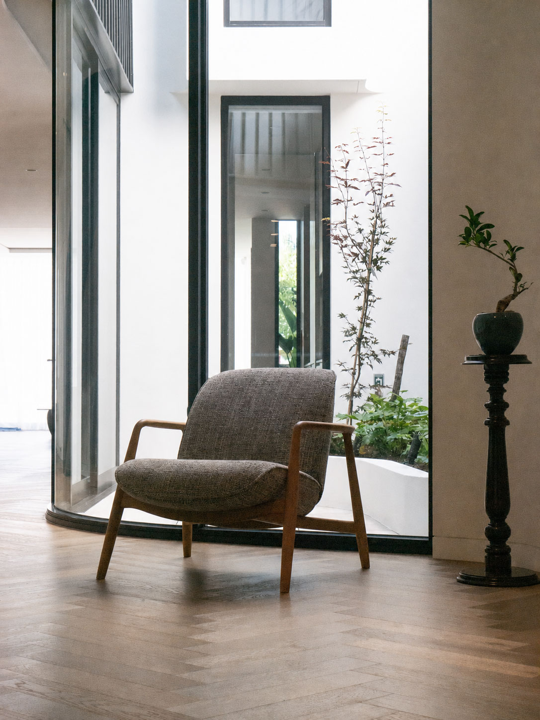Sculptural lounge chair beside curved glazing overlooking a private courtyard with herringbone timber floor — interior architecture by Mike & Morlly Architects